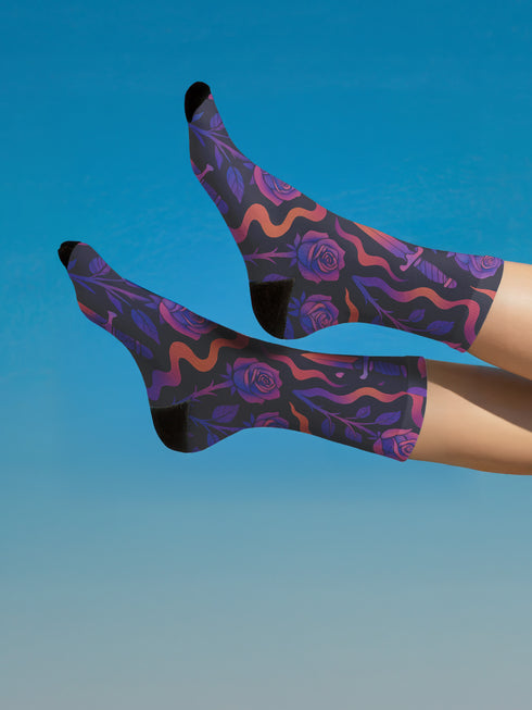 Mid-calf socks with a dark violet-black base featuring a gothic dagger and rose design in gradient shades of red, purple, and pink, with black heels and toes and an illustrated snake detail.
