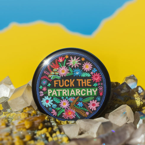 Circular black pinback button with "FUCK THE PATRIARCHY" in orange and mint green text, surrounded by embroidery-style flowers in pink, blue, and red with green foliage and blue border. Folk art feminist protest pin with glossy finish and safety pin backing in 2" and 3" sizes.