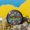 Circular black pinback button with "FUCK THE PATRIARCHY" in orange and mint green text, surrounded by embroidery-style flowers in pink, blue, and red with green foliage and blue border. Folk art feminist protest pin with glossy finish and safety pin backing in 2" and 3" sizes.