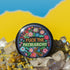 Circular black pinback button with "FUCK THE PATRIARCHY" in orange and mint green text, surrounded by embroidery-style flowers in pink, blue, and red with green foliage and blue border. Folk art feminist protest pin with glossy finish and safety pin backing in 2" and 3" sizes.