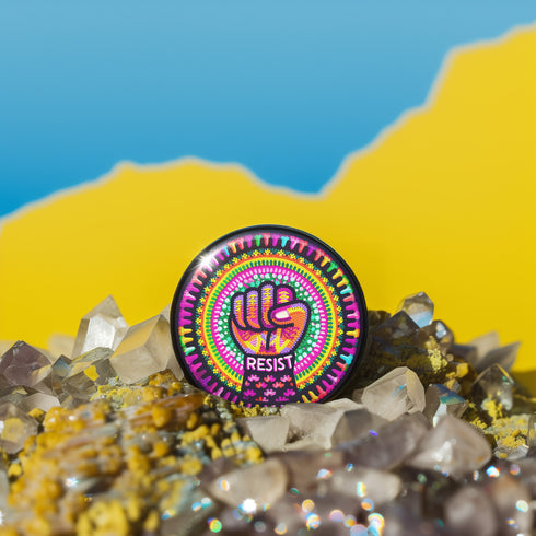 Round pinback button with psychedelic rainbow design featuring a raised black fist surrounded by concentric circles in vibrant pink, orange, green, and yellow. The word "RESIST" appears in white text below the fist, set against black background with flower details. Activist protest pin with glossy finish and safety pin backing.