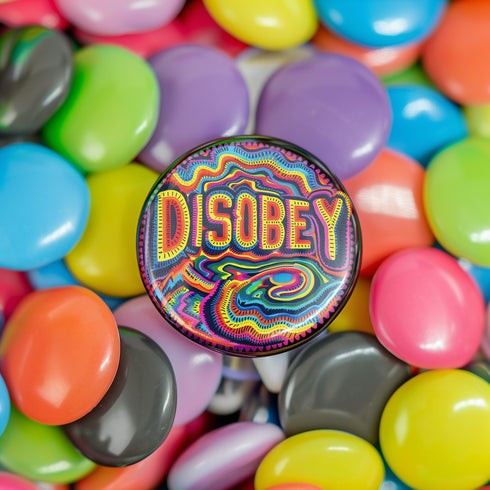 Round black pinback button with neon embroidery-style waves surrounding bold yellow and red "DISOBEY" text.