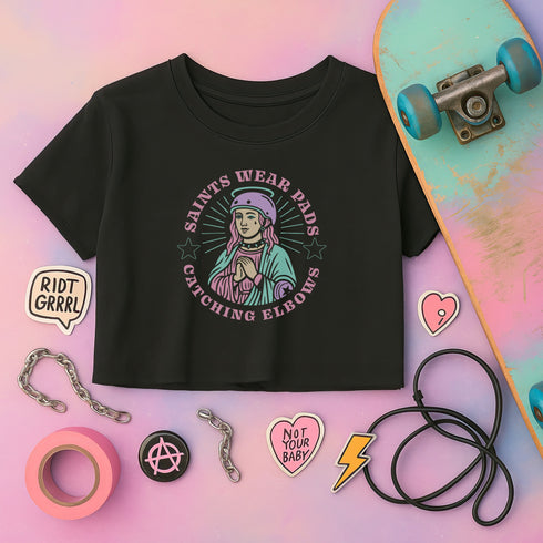 Black cropped t-shirt featuring a colorful, stylized saint figure with pink hair, wearing a helmet and pads. Surrounded by radiant lines and the phrase “Saints Wear Pads Catching Elbows” in bold pastel text. The design has a vintage punk-meets-holy aesthetic with star accents and a retro vibe. Ideal for roller skate girls, derby athletes, and goth skater fans. Available in sizes XS–2XL with raw hem details.