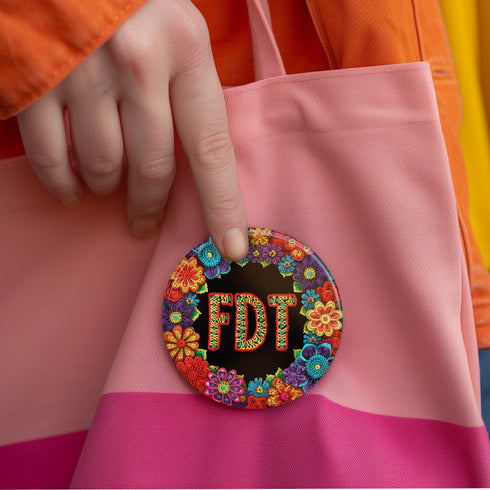 Circular black pinback button with bright 'FDT' lettering in orange/teal surrounded by colorful embroidery-style flowers in red, purple, blue, and yellow. Metal safety pin backing visible on reverse side. Political protest pin with vibrant Mexican folk art-inspired floral design.