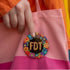 Circular black pinback button with bright 'FDT' lettering in orange/teal surrounded by colorful embroidery-style flowers in red, purple, blue, and yellow. Metal safety pin backing visible on reverse side. Political protest pin with vibrant Mexican folk art-inspired floral design.