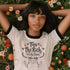 Off-white ringer t-shirt with black contrast trim on the collar and sleeve cuffs. The front features a hand-lettered design with the phrase "Tax the Rich, Feed the Poor, Pet the Cats", arranged in an elegant, whimsical script. Surrounding the text are small decorative elements, including hearts, stars, and delicate floral accents. Below the text, a simple, happy cat illustration with raised paws adds a playful and charming touch. The t-shirt has a vintage-inspired fit, made from soft cotton fabric with a sl