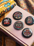 A set of five round, glossy black pinback buttons with embroidered folk art designs and bold activist slogans. Each pin features a different phrase such as "Eat the Rich" and "Resist," surrounded by colorful floral motifs.