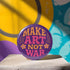 A round, glossy purple pinback button with the words "MAKE ART NOT WAR" in a groovy, retro 70s-style font. The word "MAKE" is in orange, "ART" is in a vibrant pink, and "NOT WAR" is in orange. The text is surrounded by small, stylized flower and star details. This psychedelic protest pin is a piece of activist merch with a strong metal back and safety pin clasp, perfect as an anti-war or social justice accessory for a jean jacket or backpack. The pin has a handmade, indie aesthetic.