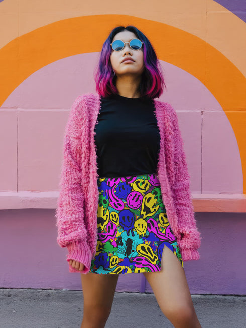 Vibrant mini skirt featuring an eye-popping psychedelic print of melting smiley faces and skulls in neon pink, bright yellow, electric blue, and lime green. The skirt has a distinctive side slit and sits above knee-length with a figure-hugging silhouette. Shown styled with various tops including a black crochet halter, black bustier, and purple blazer against colorful graffiti backdrops. The bold pattern makes this an unmissable statement piece with serious punk-meets-pop-art attitude.