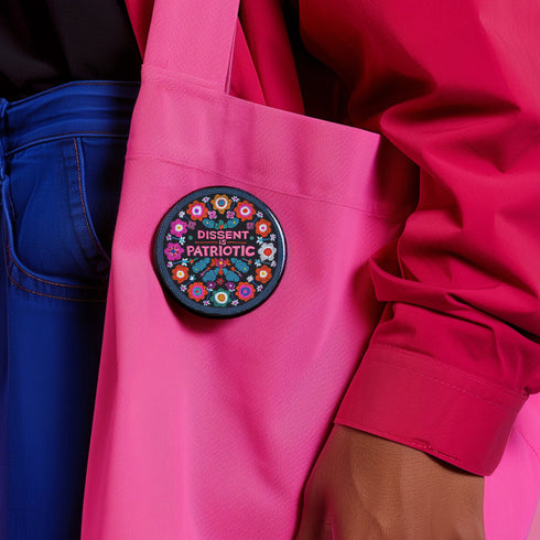 Round black pinback button with vibrant floral embroidery design surrounding bold pink and red text reading "Dissent is Patriotic."