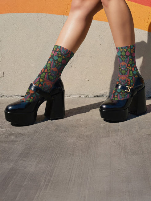 Colorful mid-calf socks with a black background featuring a whimsical folk-art bat pattern in rainbow tones of pink, orange, teal, and green, paired with matching heels and toes.