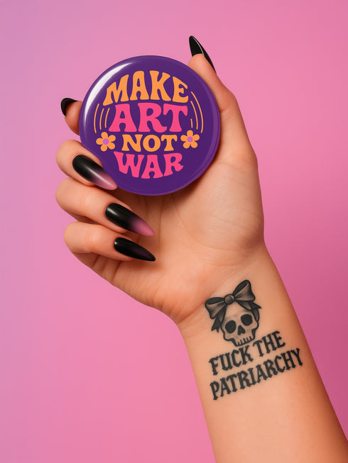 A round, glossy purple pinback button with the words "MAKE ART NOT WAR" in a groovy, retro 70s-style font. The word "MAKE" is in orange, "ART" is in a vibrant pink, and "NOT WAR" is in orange. The text is surrounded by small, stylized flower and star details. This psychedelic protest pin is a piece of activist merch with a strong metal back and safety pin clasp, perfect as an anti-war or social justice accessory for a jean jacket or backpack. The pin has a handmade, indie aesthetic.