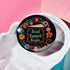 Round black pinback button with "Read Banned Books" in gold lettering on an open book image, surrounded by vibrant embroidery-style flowers in pink, orange, blue and green. Folk art-inspired literary protest pin with glossy finish and safety pin backing, available in 2" and 3" sizes.
