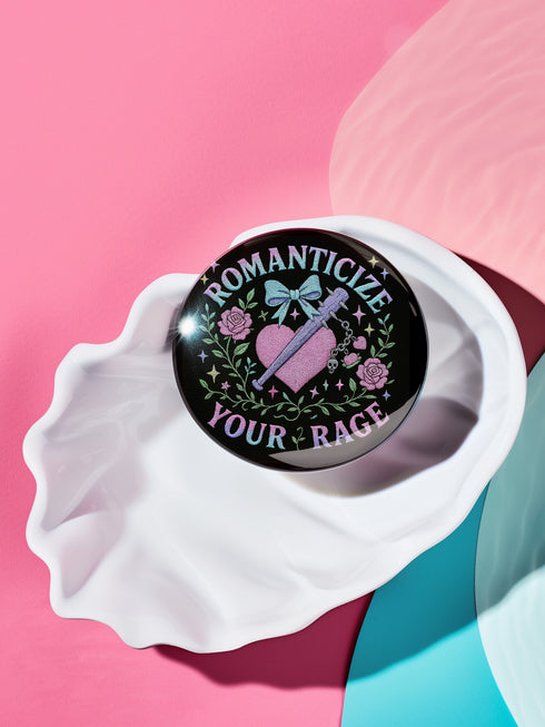 Glossy black pinback button featuring pastel gothic artwork with the phrase "Romanticize Your Rage" in bold white and lavender lettering. The design includes a pink glitter heart, a silver dagger, mint green bow, roses, stars, and delicate vines. Styled against a pink and purple gradient background, this round button blends soft romantic visuals with fierce messaging, perfect for pastel goth, feminist, or alternative fashion aesthetics.