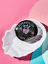 Glossy black pinback button featuring pastel gothic artwork with the phrase "Romanticize Your Rage" in bold white and lavender lettering. The design includes a pink glitter heart, a silver dagger, mint green bow, roses, stars, and delicate vines. Styled against a pink and purple gradient background, this round button blends soft romantic visuals with fierce messaging, perfect for pastel goth, feminist, or alternative fashion aesthetics.