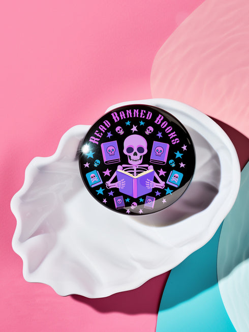 Literary rebel's badge of honor. Black circular button featuring a neon purple skeleton surrounded by floating books and stars. The text 'READ BANNED BOOKS' arcs across the top in bold purple lettering. Perfect for book-loving misfits who devour forbidden knowledge.