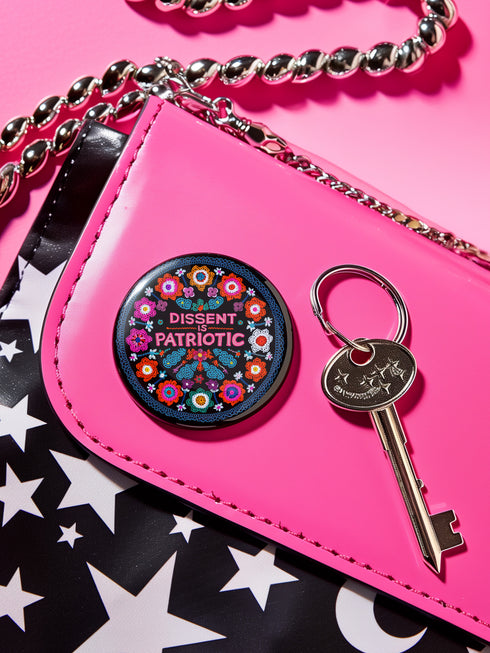 Round black pinback button with vibrant floral embroidery design surrounding bold pink and red text reading "Dissent is Patriotic."
