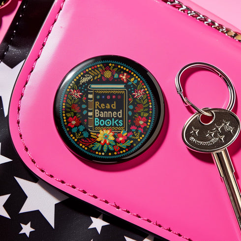 Round black pinback button with vibrant Mexican folk art-inspired embroidery design featuring red, yellow, blue and teal flowers surrounding a gold book spine with "Read Banned Books" text. Literary protest accessory with glossy finish and safety pin backing, available in 2" and 3" sizes.
