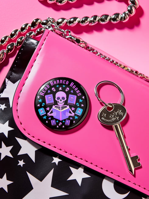 Literary rebel's badge of honor. Black circular button featuring a neon purple skeleton surrounded by floating books and stars. The text 'READ BANNED BOOKS' arcs across the top in bold purple lettering. Perfect for book-loving misfits who devour forbidden knowledge.