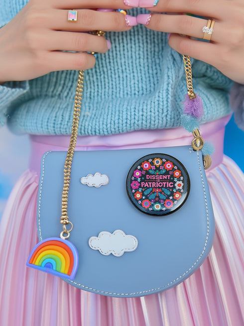 Round black pinback button with vibrant floral embroidery design surrounding bold pink and red text reading "Dissent is Patriotic."