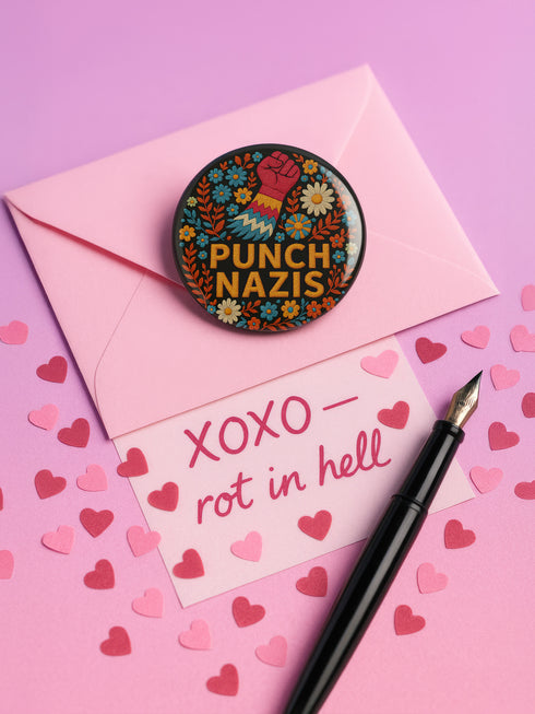 Round black pinback button with bold embroidered-style design featuring a raised red fist, zigzag sleeve, and colorful folk art flowers in orange, teal, yellow, and white. Center text reads “PUNCH NAZIS” in bright golden thread-style lettering. The design is bold, anti-fascist, and maximalist, shown on a glossy pin surface against a pink and purple gradient background. Statement accessory for protest fashion, punk style, and political activism.