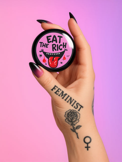 Round pink pinback button featuring a cartoon-style open mouth with sharp teeth, a red tongue, and dripping red details. The bold black text reads “EAT THE RICH” in a hand-drawn style. Glossy black rim with a vibrant, rebellious graphic design.