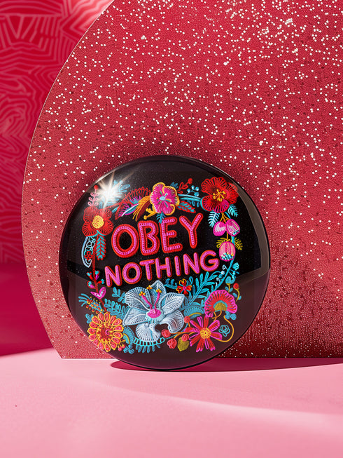 A black round pinback button featuring floral embroidery-style designs and bold red text that reads "Obey Nothing."