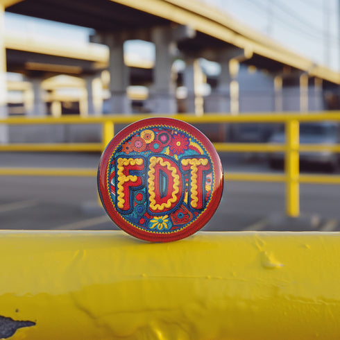 Round pinback metal button featuring an embroidered-style design on a vibrant, multicolored background. The bold, textured letters "FDT" are prominently displayed in red with intricate golden-yellow stitching details, outlined in dark blue. Surrounding the text is an ornate floral pattern with embroidered-style flowers in shades of red, orange, blue, and yellow, densely arranged for a rich, decorative effect. The outer edge is bordered with a zigzag stitching effect in red and blue. The button's back is sil