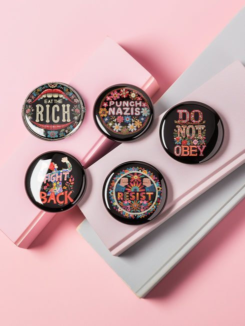 A set of five round, glossy black pinback buttons with embroidered folk art designs and bold activist slogans. Each pin features a different phrase such as "Eat the Rich" and "Resist," surrounded by colorful floral motifs.