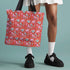 Tote bag with black handles featuring a vibrant red background covered in pastel pink and blue illustrations of skulls, roller skates, eyes, and geometric shapes. Displayed on an orange surface with dramatic shadows.