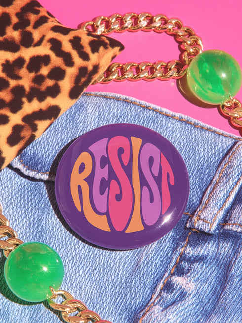 Close-up of a glossy, round purple pinback button. The button features the word "RESIST" in a bold, 70s-style psychedelic font that fills the entire surface. The bubbly, wavy letters alternate in color between a vibrant orange, a light peach-pink, and a deep magenta, creating a powerful and retro statement piece. The design has a distinct groovy and feminist protest aesthetic, making it a perfect accessory for jackets, bags, or activist apparel.