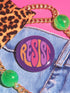 Close-up of a glossy, round purple pinback button. The button features the word "RESIST" in a bold, 70s-style psychedelic font that fills the entire surface. The bubbly, wavy letters alternate in color between a vibrant orange, a light peach-pink, and a deep magenta, creating a powerful and retro statement piece. The design has a distinct groovy and feminist protest aesthetic, making it a perfect accessory for jackets, bags, or activist apparel.