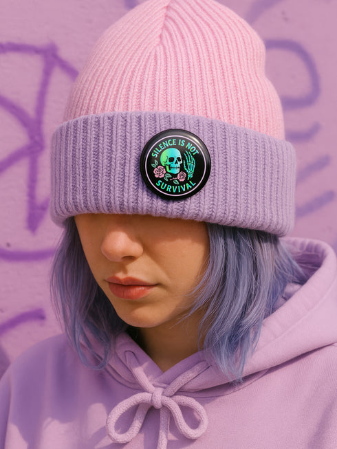 Round enamel pinback button available in 2.5″ or 3″ sizes with a black base and high-gloss finish. The design features neon green and blue gradient skull in the center, pastel pink roses and green leaves below, and mint skeleton hand on the right side. Arched text reads “SILENCE IS NOT SURVIVAL” in teal block letters. Shown styled on a denim jacket, held in hand against bright yellow backdrop, placed in a candy heart flat lay, and propped on pavement in front of graffiti wall.