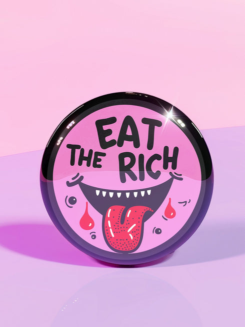 Round pink pinback button featuring a cartoon-style open mouth with sharp teeth, a red tongue, and dripping red details. The bold black text reads “EAT THE RICH” in a hand-drawn style. Glossy black rim with a vibrant, rebellious graphic design.
