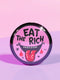 Round pink pinback button featuring a cartoon-style open mouth with sharp teeth, a red tongue, and dripping red details. The bold black text reads “EAT THE RICH” in a hand-drawn style. Glossy black rim with a vibrant, rebellious graphic design.