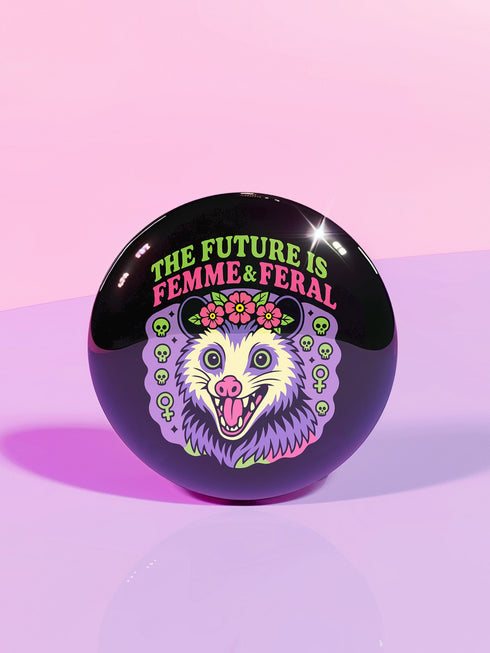 A round, black pinback button with a high-gloss finish features the text "THE FUTURE IS FEMME & FERAL" in a retro font of lime green and pastel pink. In the center, a screaming possum with wild eyes and a pink-lined mouth is illustrated in a pastel goth style with purple, yellow, and pink fur. The possum wears a pink flower crown. The illustration is framed by a circle of small skulls and pink Venus (female) symbols on a purple background. This is a piece of feminist possumcore flair.