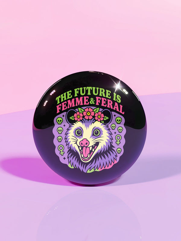 A round, black pinback button with a high-gloss finish features the text "THE FUTURE IS FEMME & FERAL" in a retro font of lime green and pastel pink. In the center, a screaming possum with wild eyes and a pink-lined mouth is illustrated in a pastel goth style with purple, yellow, and pink fur. The possum wears a pink flower crown. The illustration is framed by a circle of small skulls and pink Venus (female) symbols on a purple background. This is a piece of feminist possumcore flair.