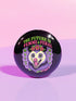 A round, black pinback button with a high-gloss finish features the text "THE FUTURE IS FEMME & FERAL" in a retro font of lime green and pastel pink. In the center, a screaming possum with wild eyes and a pink-lined mouth is illustrated in a pastel goth style with purple, yellow, and pink fur. The possum wears a pink flower crown. The illustration is framed by a circle of small skulls and pink Venus (female) symbols on a purple background. This is a piece of feminist possumcore flair.