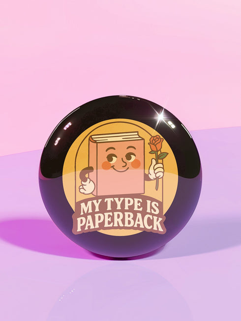 A glossy black round pin-back button with a retro 1970s-style design. The center shows a smiling cartoon paperback book holding a red rose, colored light brown with white gloves and feet. Below, “MY TYPE IS PAPERBACK” appears in chunky brown-and-tan 70s lettering. This bookish flair piece has a high-gloss, scratch-resistant mylar finish, secure safety pin backing, and comes in two- or three-inch sizes.