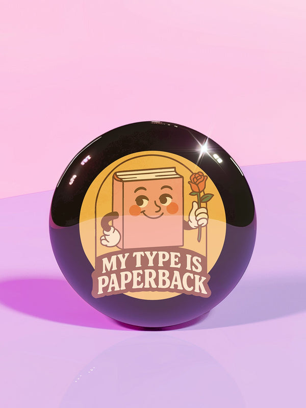 A glossy black round pin-back button with a retro 1970s-style design. The center shows a smiling cartoon paperback book holding a red rose, colored light brown with white gloves and feet. Below, “MY TYPE IS PAPERBACK” appears in chunky brown-and-tan 70s lettering. This bookish flair piece has a high-gloss, scratch-resistant mylar finish, secure safety pin backing, and comes in two- or three-inch sizes.