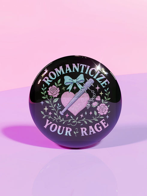 Glossy black pinback button featuring pastel gothic artwork with the phrase "Romanticize Your Rage" in bold white and lavender lettering. The design includes a pink glitter heart, a silver dagger, mint green bow, roses, stars, and delicate vines. Styled against a pink and purple gradient background, this round button blends soft romantic visuals with fierce messaging, perfect for pastel goth, feminist, or alternative fashion aesthetics.