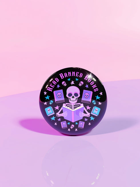 Literary rebel's badge of honor. Black circular button featuring a neon purple skeleton surrounded by floating books and stars. The text 'READ BANNED BOOKS' arcs across the top in bold purple lettering. Perfect for book-loving misfits who devour forbidden knowledge.