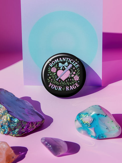Glossy black pinback button featuring pastel gothic artwork with the phrase "Romanticize Your Rage" in bold white and lavender lettering. The design includes a pink glitter heart, a silver dagger, mint green bow, roses, stars, and delicate vines. Styled against a pink and purple gradient background, this round button blends soft romantic visuals with fierce messaging, perfect for pastel goth, feminist, or alternative fashion aesthetics.