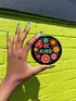 Bold "BE KIND" pinback button featuring vibrant psychedelic flowers in hot pink, teal, yellow, and orange on black background. Retro 70s-inspired typography in coral and mint green surrounded by colorful blooms. Statement activist accessory perfect for punk fashion, protest wear, and battle vests. Glossy UV-resistant finish with safety pin backing available in 2" and 3" sizes.