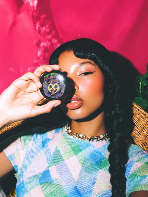 A round, black pinback button with a high-gloss finish features the text "THE FUTURE IS FEMME & FERAL" in a retro font of lime green and pastel pink. In the center, a screaming possum with wild eyes and a pink-lined mouth is illustrated in a pastel goth style with purple, yellow, and pink fur. The possum wears a pink flower crown. The illustration is framed by a circle of small skulls and pink Venus (female) symbols on a purple background. This is a piece of feminist possumcore flair.