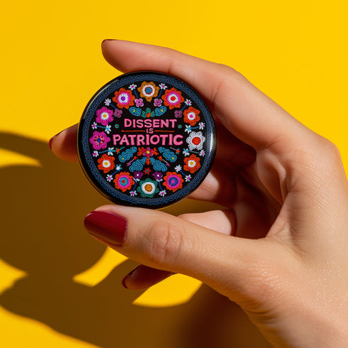 Round black pinback button with vibrant floral embroidery design surrounding bold pink and red text reading "Dissent is Patriotic."