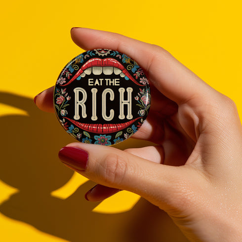 A round pin-back button with a bold, embroidered-style design featuring the phrase "EAT THE RICH" in large, vintage-style lettering. The typography is framed by a set of open red lips with detailed stitching, revealing white teeth. Surrounding the mouth are intricate floral patterns in shades of blue, red, and green, adding a decorative, folk-art-inspired touch. The button has a black background, making the details and lettering stand out
