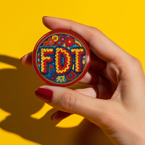 Round pinback metal button featuring an embroidered-style design on a vibrant, multicolored background. The bold, textured letters "FDT" are prominently displayed in red with intricate golden-yellow stitching details, outlined in dark blue. Surrounding the text is an ornate floral pattern with embroidered-style flowers in shades of red, orange, blue, and yellow, densely arranged for a rich, decorative effect. The outer edge is bordered with a zigzag stitching effect in red and blue. The button's back is sil