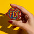 Round pinback metal button featuring an embroidered-style design on a vibrant, multicolored background. The bold, textured letters "FDT" are prominently displayed in red with intricate golden-yellow stitching details, outlined in dark blue. Surrounding the text is an ornate floral pattern with embroidered-style flowers in shades of red, orange, blue, and yellow, densely arranged for a rich, decorative effect. The outer edge is bordered with a zigzag stitching effect in red and blue. The button's back is sil