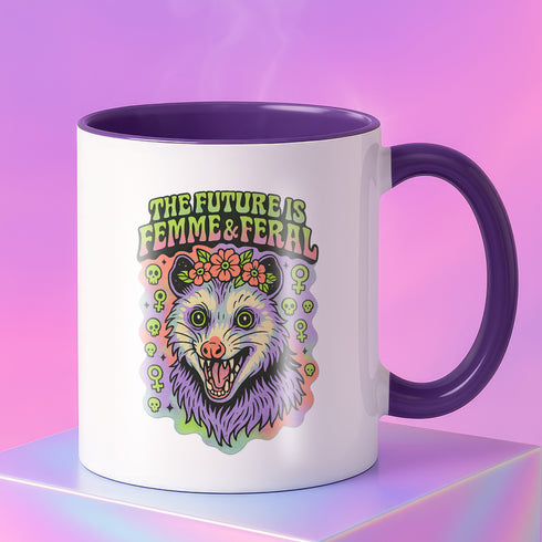 White ceramic coffee mug with pink or orange interior and handle, featuring a double-sided colorful design of a screaming possum wearing a flower crown with the phrase “The Future is Femme & Feral.” The design includes skulls and feminist symbols with a neon, pastel, and spooky aesthetic. Available in 11oz and 15oz.